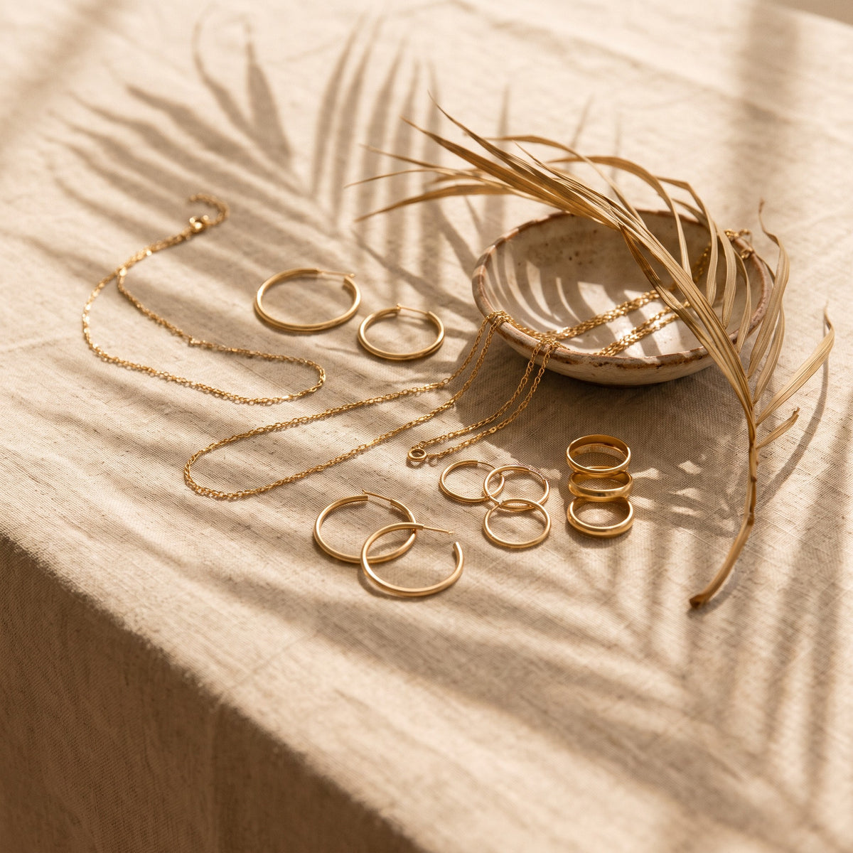 Gold chains, hoops and stacked rings on sunlit linen