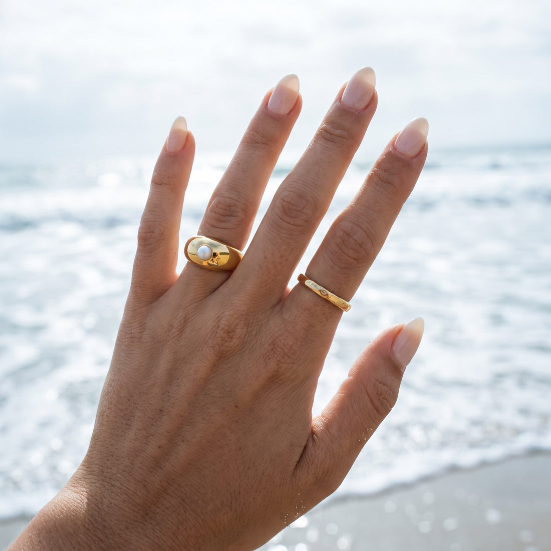 Hand wearing a gold dome ring with a pearl and a slim gold band by the sea