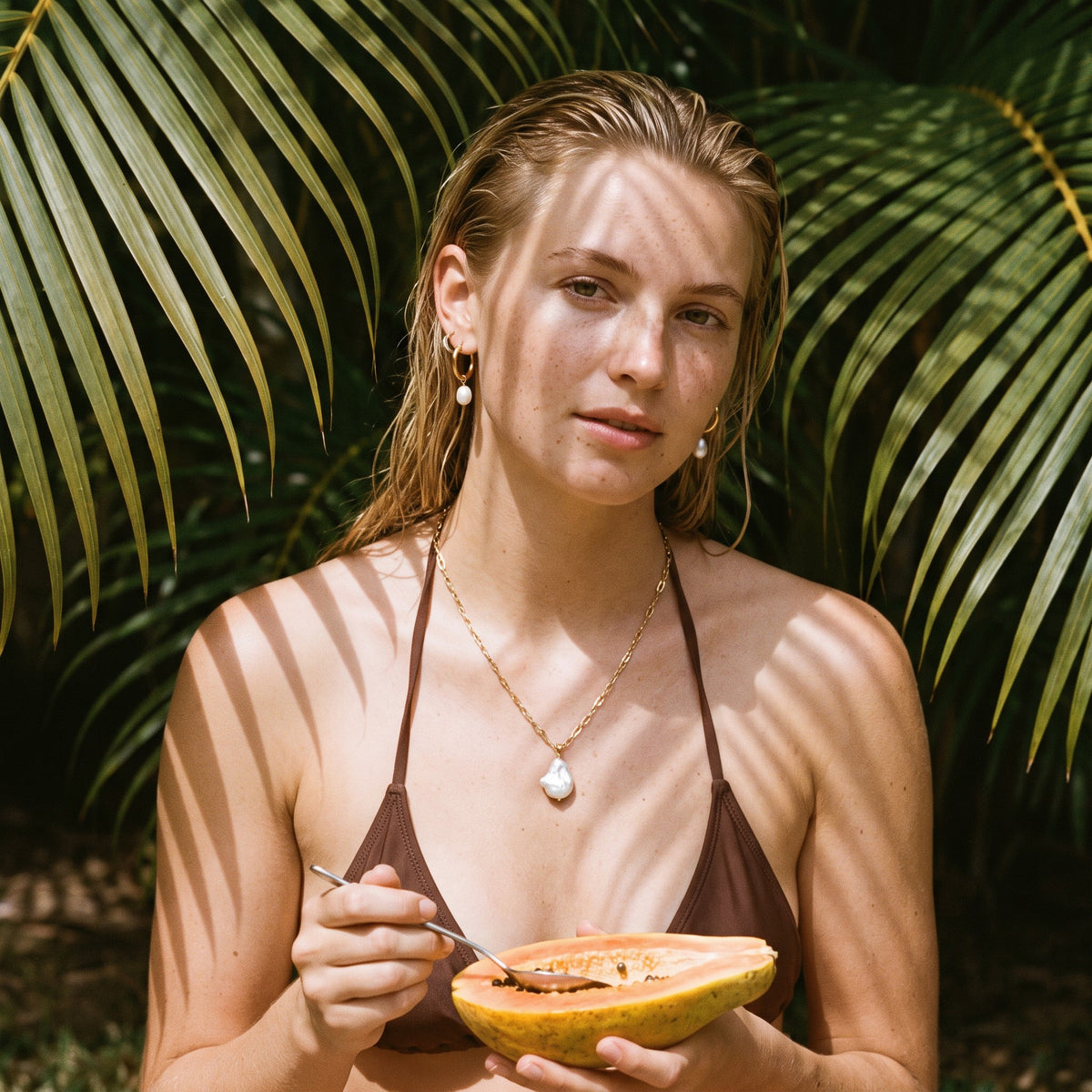 Blonde woman under palm leaves wearing a pearl pendant and pearl drop hoops